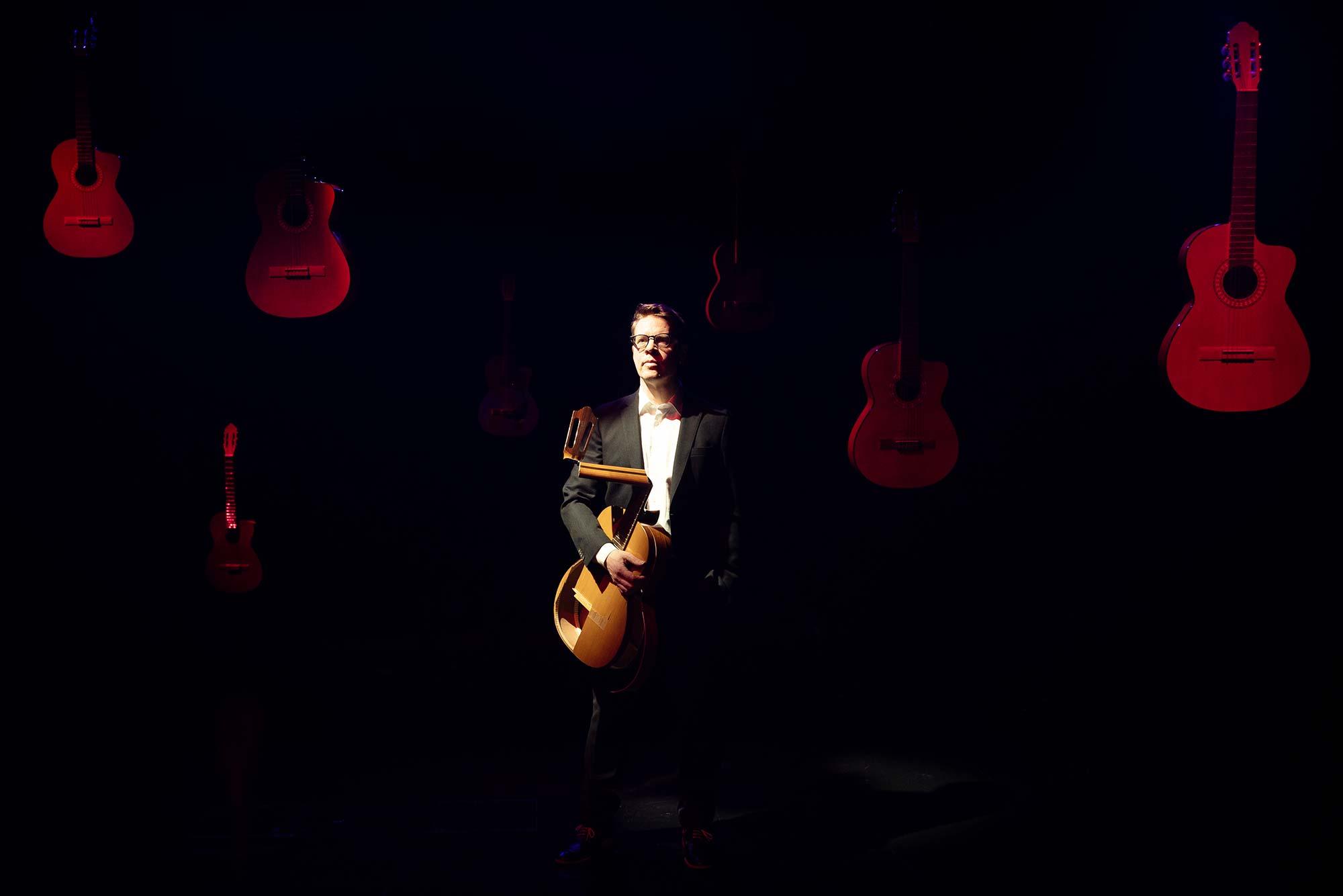 Keskellä Petri Kumela kädessään rikkinäinen kitara. Tumma tausta, jossa punaisella valolla valaistuja ehjiä kitaroita.