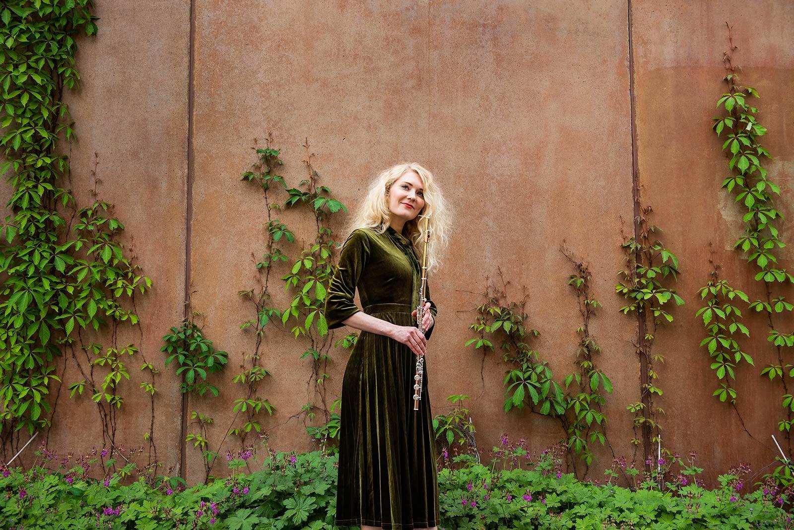 Johanna Suvenmetsä huilun kanssa betonisen seinän edessä, jota vasten kasvaa vihreitä köynnöksiä.